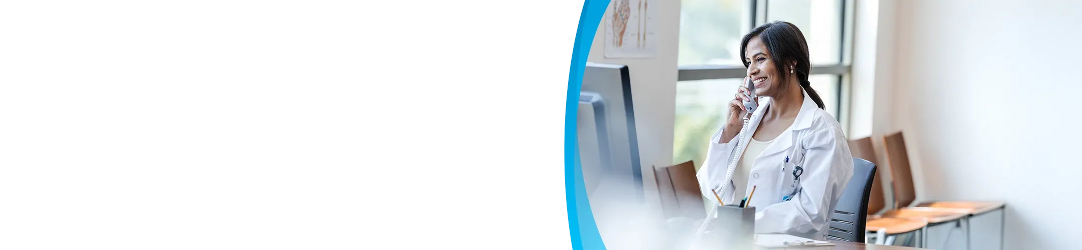 A smiling female doctor or medical professional wearing a white lab coat is sitting at a desk and talking on a landline phone next to a computer monitor.